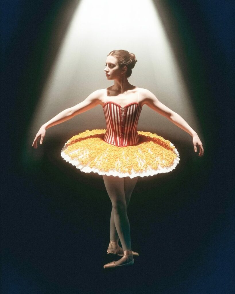 Una bailarina en pose elegante, en un fondo blanco minimalista, con un atuendo inspirado en una caja de palomitas de cine.